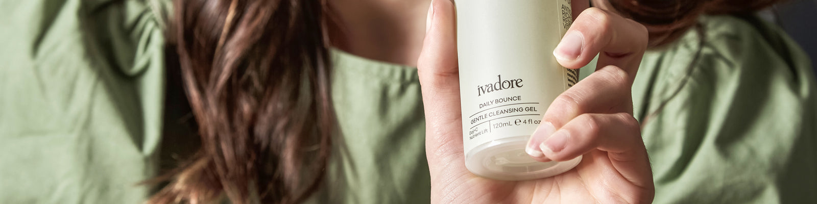 Image shows woman with brown hair in olive green dress holding a bottle of Ivadore Daily Bounce Cleansing Gel